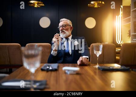 Homme senior à cheveux gris tenant et fume e-cigarette dans un restaurant riche. IQOS, technologies modernes, arrêter la fumée Banque D'Images