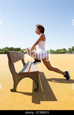 Jeune femme s'étirant sur un banc extérieur. Elle porte un short et un débardeur. Banque D'Images