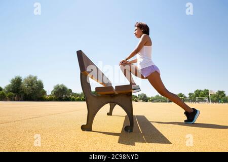 Jeune femme qui étire ses muscles sur un banc d'extérieur avant de faire du sport. Espace pour le texte. Banque D'Images