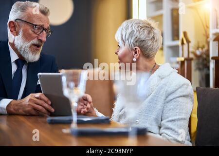 Joyeux, joyeux, heureux, couple mature amoureux de passer du temps au restaurant tout en discutant et regardant les uns les autres. Un homme âgé montre quelque chose sur une tablette Banque D'Images