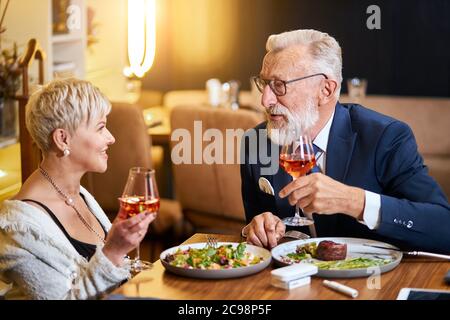 Un couple senior élégant et élégant se regarde les uns les autres et discute, entretient des relations étroites. Un homme aux cheveux gris en tuxedo lève un verre. Utilisez une cigarette électronique, un singe. Mo Banque D'Images