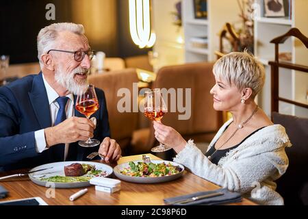 Un couple senior élégant et élégant se regarde les uns les autres et discute, entretient des relations étroites. Un homme aux cheveux gris en tuxedo lève un verre. Utilisez une cigarette électronique, un singe. Mo Banque D'Images