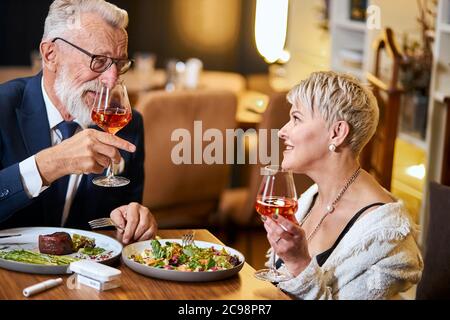 Un couple senior élégant et élégant se regarde les uns les autres et discute, entretient des relations étroites. Un homme aux cheveux gris en tuxedo lève un verre. Utilisez une cigarette électronique, un singe. Mo Banque D'Images