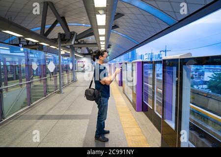 Province de Nonthaburi, Thaïlande - 14 juillet 2020, un homme asiatique se tient debout et joue son téléphone mobile en attendant le train sur la plate-forme mauve alo Banque D'Images