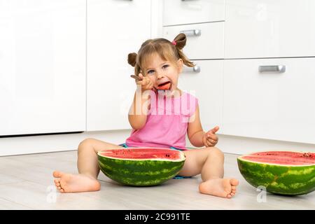 Une petite fille mignonne est assise sur un sol et mange un melon d'eau avec une cuillère Banque D'Images