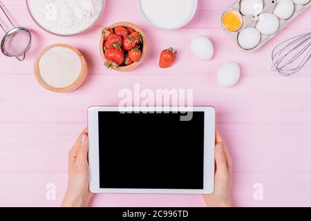 Les femmes tiennent une tablette numérique avec écran vierge parmi les ingrédients pour cuire un gâteau de fraise sur une table rose, vue de dessus. Banque D'Images