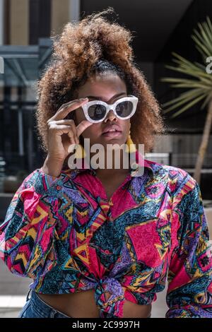 Femme afro-américaine dans une tenue à la mode et des lunettes de soleil debout en ville le jour ensoleillé en été Banque D'Images