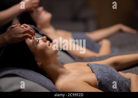 deux belles femmes caucasiennes reçoivent un massage facial, les jeunes femmes s'étendent sur le lit tandis que les masseurs touchent la peau de la tête et du visage, font le massage avec soin Banque D'Images