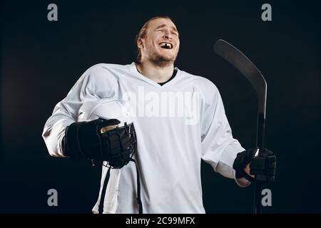 Heureux beau joueur de hockey avec une dent de devant cassée riant de caméra, debout avec stick en uniforme blanc, isolé sur fond noir Banque D'Images