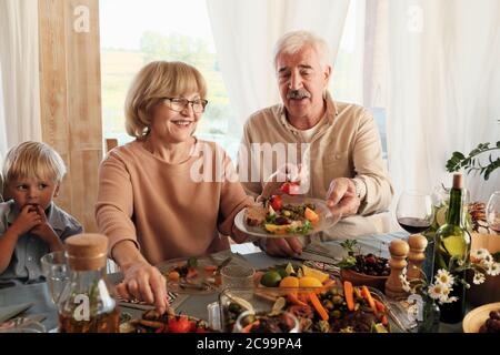 Bonne femme âgée mettant des légumes sur l'assiette de son mari pendant qu'ils sont assis à la table pendant le dîner de vacances Banque D'Images