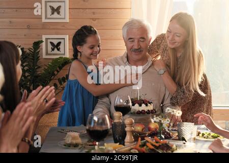 Homme âgé assis à la table avec un gâteau d'anniversaire tandis que ses petites-filles le félicitant de son anniversaire à la maison Banque D'Images