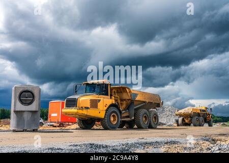 Deux poids lourds à benne jaune industrielle stationnés près du chantier de construction avec des matériaux de construction en stock, des nuages bleu foncé avant la pluie. Été ensoleillé Banque D'Images