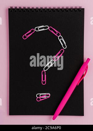 Point d'interrogation composé de clips sur bloc-notes noir vierge sur fond rose. Pose à plat. Banque D'Images