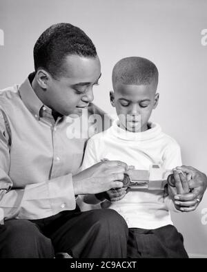 ANNÉES 1960 HOMME AFRO-AMÉRICAIN PÈRE AVEC SON FILS ASSIS CÔTE-À-CÔTE TENANT VOITURE JOUET - N2230 HAR001 HARS COMMUNICATION JEUNES ADULTES TRAVAIL D'ÉQUIPE FILS FAMILLES JOIE STYLE DE VIE SATISFACTION STUDIO TOURNÉ MAISON VIE COPIE ESPACE AMITIÉ DEMI-LONGUEUR PERSONNES INSPIRATION HOMMES PÈRES B&W BONHEUR DÉCOUVERTE EXPLIQUANT SON AFRO-AMÉRICAIN AFRO-AMÉRICAIN DADS NOIR SOUTIEN ETHNIQUE CROISSANCE JUVÉNILES CÔTE À CÔTE ENSEMBLE JEUNE HOMME ADULTE NOIR ET BLANC HAR001 AFRO-AMÉRICAINS À L'ANCIENNE Banque D'Images