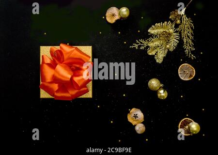 Boîte cadeau ornée d'un noeud rouge et de boules dorées. Fond noir décoré avec des étoiles confetti. Flat lay, vue de dessus. Concept de Noël. Banque D'Images