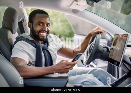 Heureux souriant Africain américain conducteur assis derrière le volant de direction autonome d'une voiture électrique moderne autonome. Un homme heureux tient le téléphone Banque D'Images