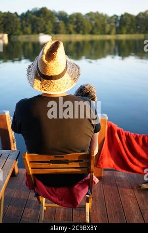 Arrière de l'homme avec chapeau de paille et chien assis sur une chaise en bois regardant le lac calme au coucher du soleil Banque D'Images