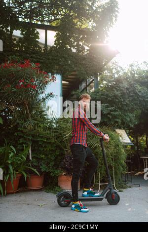 Vue latérale d'un jeune homme caucasien à barbe avec lunettes de soleil promenades sur la rue e scooter Banque D'Images