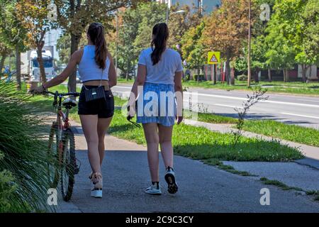 Zrenjanin, Serbie, 29 juillet 2020. Deux belles filles marchent autour de la ville détendu et parle. L'un d'eux pousse une bicyclette à côté d'elle. Banque D'Images