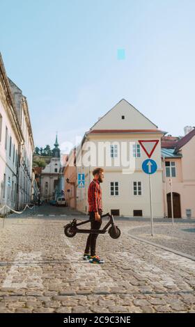 Un beau jeune homme caucasien avec une barbe et des lunettes de soleil se promène au-dessus d'un piéton, tenant un scooter électrique dans la main Banque D'Images