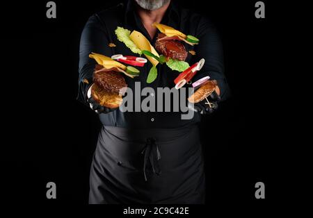 chef dans une chemise noire, un tablier et des gants noirs de latex se tient sur un fond noir, dans ses mains volant ingrédients de cheeseburger: un pain avec des graines de sésame Banque D'Images