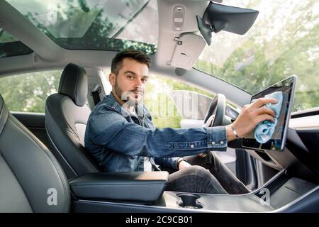 Portrait d'un jeune homme caucasien souriant, avec une barbe assise à l'intérieur de sa voiture moderne à direction automatique et essuyant la poussière de l'écran tactile avec Banque D'Images