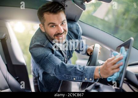 Gros plan portrait d'un beau caucasien souriant avec barbe essuyant la poussière de l'écran tactile avec un chiffon en microfibre, à l'intérieur de son art de l'auto moderne Banque D'Images