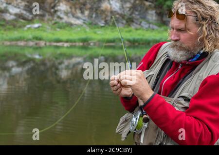 Pêche à la mouche mature pêcheur dans des verres polarisants nouant la mouche du crochet de pêche sur la canne à pêche Banque D'Images