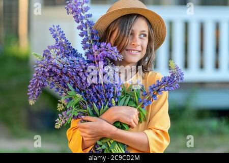 Une fille souriante tient un beau grand bouquet de lupin dans ses mains. Un enfant dans un chapeau de paille. Jour d'été ensoleillé. Banque D'Images