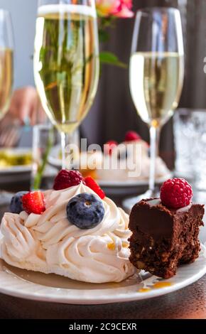 Gâteau meringue aux baies et gâteau au chocolat sur une assiette blanche avec deux verres de champagne sur fond de bokeh. Banque D'Images