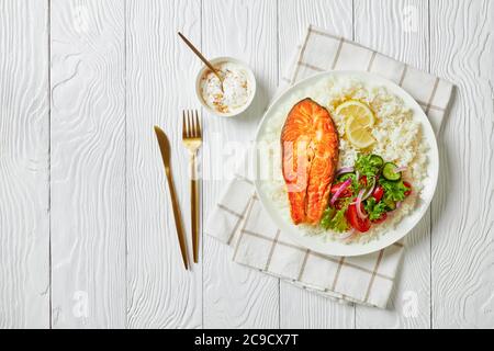 gros plan du steak de truite frit servi avec de la salade de riz et de légumes sur une assiette blanche sur une table en bois avec de la sauce moutarde au yaourt et des couverts dorés, la Banque D'Images