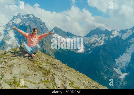 Jeune femme heureuse assise sur le rocher contre un ciel nuageux par beau soleil en montagne. La femme voyageur admire une vue magnifique dans la région montagneuse. Banque D'Images