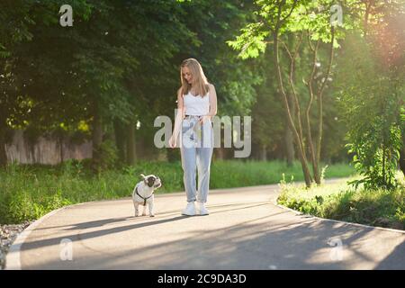 Jeune femme souriante dans un ensemble décontracté marchant avec un joli boudogue français sur la laisse, regardant le chien. Vue de face de la magnifique Happy caucasiens fille appréciant la promenade avec un animal de compagnie charmant dans le parc de la ville. Banque D'Images