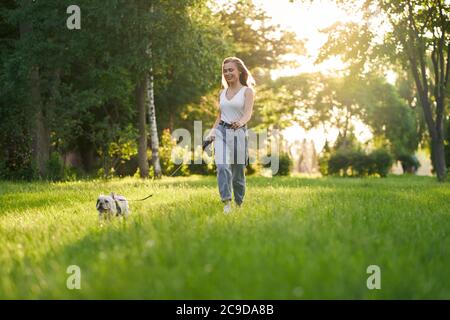 Jeune femme heureuse vêtue d'une tenue décontractée souriante et en train de courir sur l'herbe avec le boudogue français sur la laisse. Magnifique fille caucasienne appréciant le coucher du soleil d'été avec un animal de compagnie de race pure dans le parc de la ville, s'amuser à l'extérieur. Banque D'Images