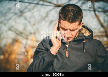 Jeune homme sérieux tenant un téléphone portable, utilisant un smartphone, faisant un appel, parlant au téléphone, debout sur une rue ensoleillée Banque D'Images