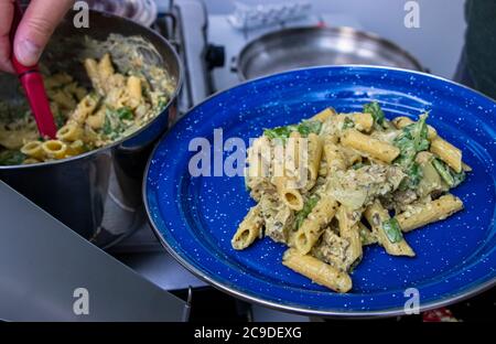 Homme servant du pesto cuit, du fromage à la crème et du poulet végétarien pâtes One Pot Penne dans une assiette en émail à côté d'un poêle Coleman, repas du camping Banque D'Images