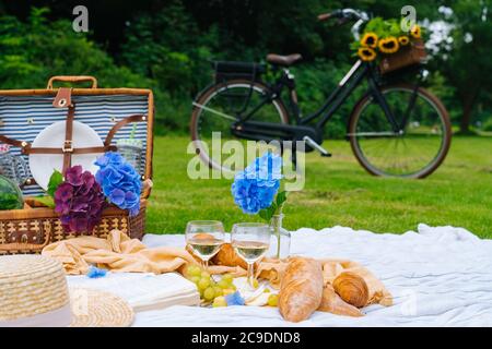 Pique-nique d'été le jour ensoleillé avec pain, fruits, bouquet de fleurs d'hortensia, verres de vin, chapeau, livre. Panier pique-nique sur l'herbe avec nourriture et boisson sur une couverture en tricot. Vélo en arrière-plan. Mise au point sélective. Banque D'Images