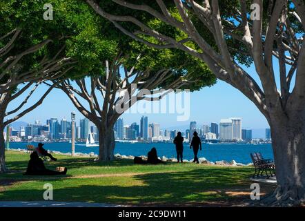 Vue sur la ville de San Diego avec bâtiments, parc et personnes en silhouette relaxante. Jour d'été ensoleillé sous les arbres ombrageant la pelouse. Banque D'Images