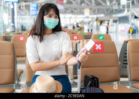 Femme asiatique touriste portant le masque regardant heureux tout en souriant derrière le masque et assis social distancer chaise dans le terminal de l'aéroport pendant la protection Banque D'Images