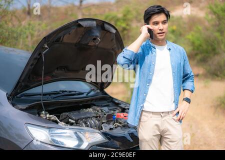 Jeune homme asiatique malcontent de parler sur un téléphone portable devant le capot ouvert cassé voiture Sur Country Road Téléphone Pour l'aide. Une Voiture Cassée Sur La Route I Banque D'Images