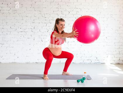 Une jeune femme enceinte s'accroupite avec une balle de fitness. Portrait pleine longueur. Banque D'Images
