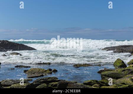 Roches vertes de mousse entourant un Rockpool avec des vagues de l'océan de couleur aqua se brisant sur de grandes roches avec ciel bleu avec des nuages en arrière-plan Banque D'Images