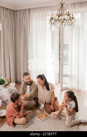 Une jeune famille heureuse et excitée, assise sur le sol dans le nouveau salon et manger de la pizza tout en discutant ensemble Banque D'Images