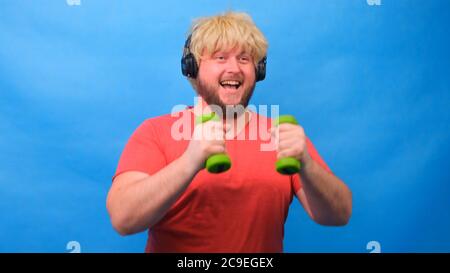 Drôle d'homme de graisse freaky dans une perruque avec des haltères faisant des exercices sur un fond bleu. Banque D'Images