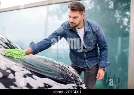 Vue latérale d'un jeune homme beau barbu dans une chemise de Jean, en lavant sa capuche de voiture avec une éponge verte et du détergent au lavage auto extérieur Banque D'Images