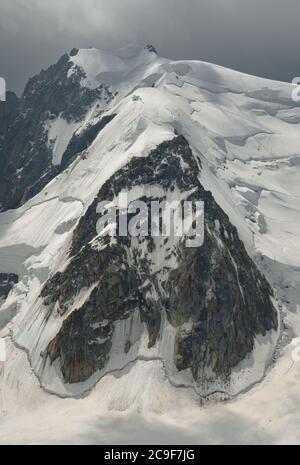 Face rocheuse de forme inhabituelle sur le côté d'une montagne enneigée. Une lumière vive brille à travers un ciel nuageux au-dessus. Composition graphique. Banque D'Images