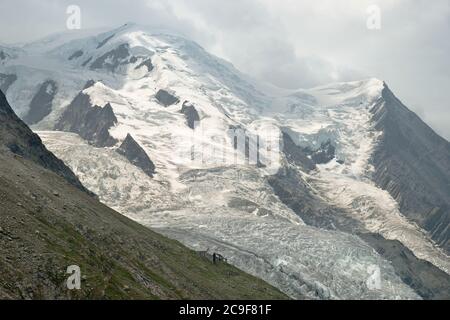 Un glacier qui traverse des montagnes enneigées. Un petit bâtiment abandonné se trouve en face de l'immense glacier et les nuages traversent la montagne. Banque D'Images