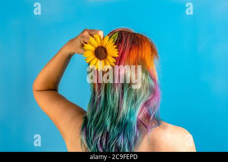 Portrait de la jeune femme aux cheveux multicolores. Vue arrière avec un joli tournesol rouge et frais à la main sur fond bleu. Banque D'Images