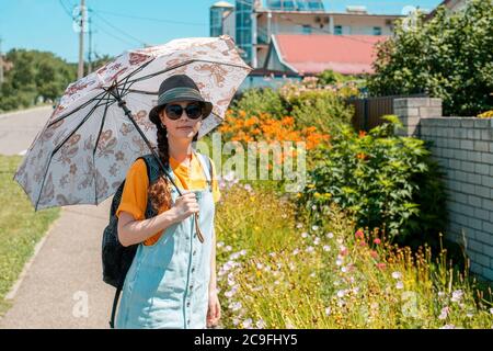 Une femme se tient sur une rue verte dans un chapeau de paille, des lunettes et un parapluie, se protégeant du soleil. Extérieur. Banque D'Images