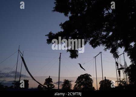 Silhoueté dans le ciel du soir, une figure interprète des acrobaties trapèze à Ruskin Park à Lambeth, le 29 juillet 2020, à Londres, en Angleterre. L'école Trapeze de TLCC arrive à Ruskin Park chaque été, bien qu'au cours de la pandémie du coronavirus, ils aient dû retarder leur présence dans ce sud de London Park. Banque D'Images